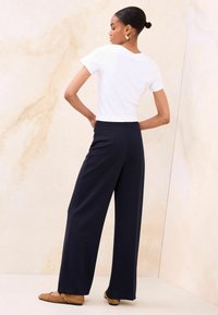 Valge lühikeste varrukatega top, kombineeritud tumeliste laia säärega pükstega, millel on elastne vöökoht, pehme ja neutraalse taustaga.