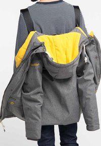 Persona che indossa una camicia a righe e porta una giacca grigia con fodera gialla sulla schiena, utilizzando le cinghie di uno zaino nero.