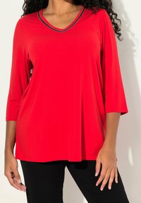 Top rosso a maniche corte con scollo a V e rifiniture metalliche lungo il collo. Realizzato in un tessuto liscio, presenta un design rilassato e fluido.