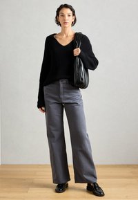 Egy nő áll, fekete kötött pulóverben, szürke, bőszárú nadrágban, fekete mokaszinban és egy nagy fekete, redőzött válltáskával. 