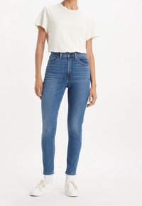 Jeans de tiro alto azul con un ajuste slim, hechos de denim. Presentan cinco bolsillos, pasadores para cinturón y un leve desgaste. Combinados con una camiseta blanca.