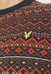 Flerfärgad stickad tröja med geometriska mönster i röd, brun, vit och marinblå, med ett litet gult fågelmärke på bröstet.