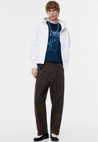 Világosszürke, rövidített kapucnis pulóver sötétkék grafikás póló felett; barna mintás, bő szárú nadrág. Fekete cipők krém színű részletekkel. Ezüst nyaklánc.