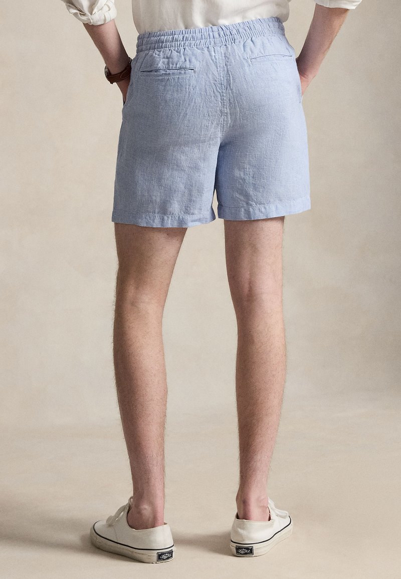 Personne portant un short bleu clair à taille élastique, des baskets blanches et une chemise blanche à manches longues avec les manches roulées, debout les mains dans les poches.