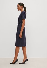 Navy-Kleid aus glattem Stoff, mit kurzen Ärmeln, tailliertem Schnitt und einem Gürteldetail; kombiniert mit schwarzen, spitzen High Heels.