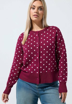 Kvinde iført en vinrød knappet cardigan med hvide prikker og blå jeans mod en ensfarvet lys baggrund.