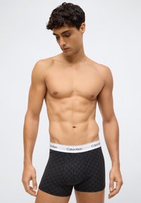 Muž bez trička s tmavými kučeravými vlasmi, ktorý má na sebe čierne vzorované boxerky Calvin Klein, stojí pred jednoduchým svetlým pozadím.