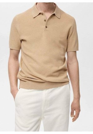 Mand iført en beige kortærmet strikket polo med tre knapper og hvide bukser, med hænderne delvist i lommerne.