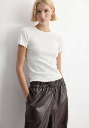 Blondínka v priliehavom bielom krátkom rukáve s cvokmi na okrajoch rukávov a tmavohnedých kožených nohaviciach s vysokým pásom.