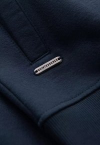 Tessuto di felpa blu navy con una texture liscia. Presenta un'etichetta metallica argentea incisa con "QUINTESSENCE" attaccata alla cucitura.