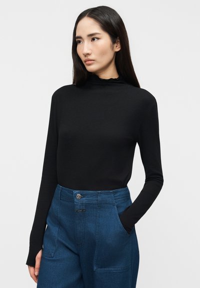 Femme aux longs cheveux noirs portant un haut noir à manches longues et un pantalon bleu taille haute, debout avec la main gauche dans la poche, sur un fond gris.