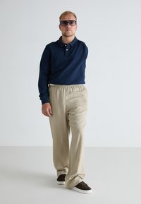 Cămașă polo de navy cu mânecă lungă, pantaloni bej cu talie elastică și adidași maro. Ochelari cu lentile mari de culoare închisă. Croială simplă, lejeră.