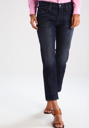 Femme portant un jean droit bleu foncé, une chemise rose à manches longues et des sandales marron à boucle, marchant sur un fond neutre.