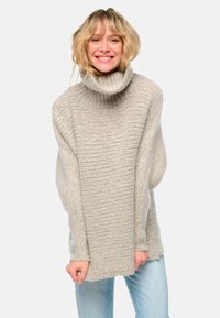 Lächelnde Frau mit kurzem blonden Haar, die einen grob gestrickten beigen Rollkragenpullover und hellblaue Jeans trägt, vor einem schlichten weißen Hintergrund.