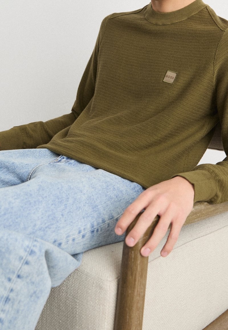 T-shirt à manches longues vert olive avec un motif texturé et une petite étiquette de marque sur la poitrine, associé à un jean en denim bleu clair.