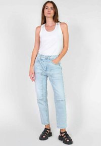 Lichtblauwe denim jeans met een relaxed fit, voorzien van voorzakken en unieke stiksels. Gecombineerd met een witte tanktop en zwarte sandalen.