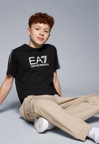 Melns īsām piedurknēm T-krekls ar baltu logo tekstu, kombinēts ar gaiši brūnām biksēm. Modelis sēž, valkājot baltas kedas. Gluda auduma, brīvs piegriezums.