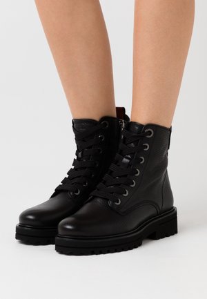 Stivaletti alla caviglia neri in pelle con lacci e suole spesse indossati su gambe nude su uno sfondo bianco semplice.