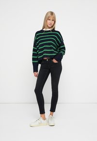 Suéter corto a rayas azul marino y verde con un corte holgado, combinado con jeans ajustados negros y zapatillas blancas. Textura suave, diseño casual.