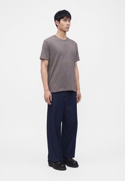 Jeune homme debout portant un t-shirt gris à manches courtes, un pantalon large bleu foncé et des chaussures noires sur un fond blanc uni.