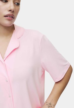 Jeune personne portant une chemise rose à manches courtes et un tatouage de constellation d'étoiles sur l'avant-bras intérieur, regardant sur le côté.