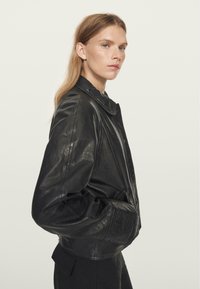 Černá kožená bunda s límcem, jemnou texturou a volným střihem. Obsahuje boční kapsy a mírně zkrácenou délku.