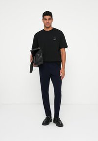 T-shirt preto de manga curta com um pequeno logótipo, combinado com calças ajustadas azul-marinho. O modelo segura uma bolsa de couro preta e usa sapatos pretos.