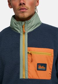 Ζακέτα από φλις σε ναυτικό μπλε με πράσινο γιακά. Διαθέτει πορτοκαλί τσέπη, κίτρινο φερμουάρ και ένα σήμα «Adventure Division» στο μπροστινό μέρος.