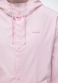 Ροζ ανοιχτόχρωμο αδιάβροχο με κουκούλα, κορδόνια ρύθμισης, κουμπιά τύπου snap κατά μήκος του εμπρός μέρους και μικρό ορθογώνιο λογότυπο "Hunter" στο στήθος.