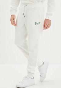 Witte trainingsbroek met een drawstring taille, voorzien van een groene "Creek's Original" logo op de dij. Zachte textuur met geribde boorden.