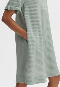 Hellgrünes gestreiftes Kleid mit lockerer Passform, kurzen umgeschlagenen Ärmeln, Knopfleiste und seitlichen Taschen. Der strukturierte Stoff verleiht subtilen Tiefeneffekt.