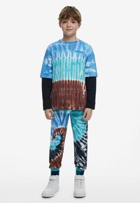 Dieťa s krátkymi hnedými vlasmi, oblečené v modrom, hnedom a čiernom tričku s efektom tie-dye a zodpovedajúcich nohaviciach, biele tenisky a stojí pred bielym pozadím.