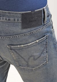 Ljusblå denimjeans med en mörk läderetikett i midjan, som har en distinkt bakficka med böjd sömnad.