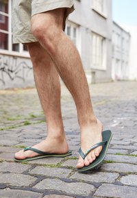 Uomo che indossa pantaloni corti beige e infradito verdi, mentre cammina su una strada ciottolata con edifici sullo sfondo.