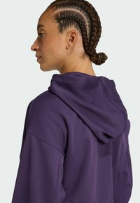 Sudadera morada con un patrón texturizado, ajuste holgado y capucha con cordón. Presenta trenzas y una vista de perfil lateral del usuario.