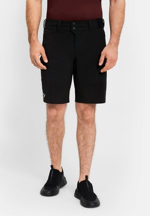Mann trägt schwarze knielange Shorts mit Vordertaschen und schwarze Slip-On-Sportschuhe, steht vor einem schlichten Hintergrund.