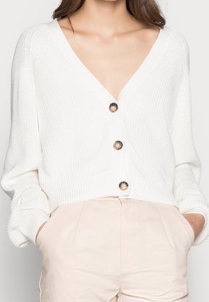 Cardigan blanc à col en V boutonné porté sur un pantalon taille haute beige clair, mains dans les poches, cadré du cou aux hanches.