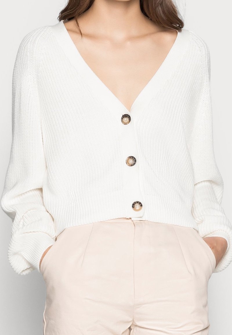 Cardigan blanc à col en V boutonné porté sur un pantalon taille haute beige clair, mains dans les poches, cadré du cou aux hanches.