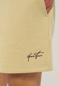 Μπεζ σορτς με μαύρο καλλιγραφικό κείμενο κοντά στο τελείωμα, το χέρι ακουμπά στην πλαϊνή τσέπη, δείχνοντας την υφή του υφάσματος και τις λεπτομέρειες της ραφής.