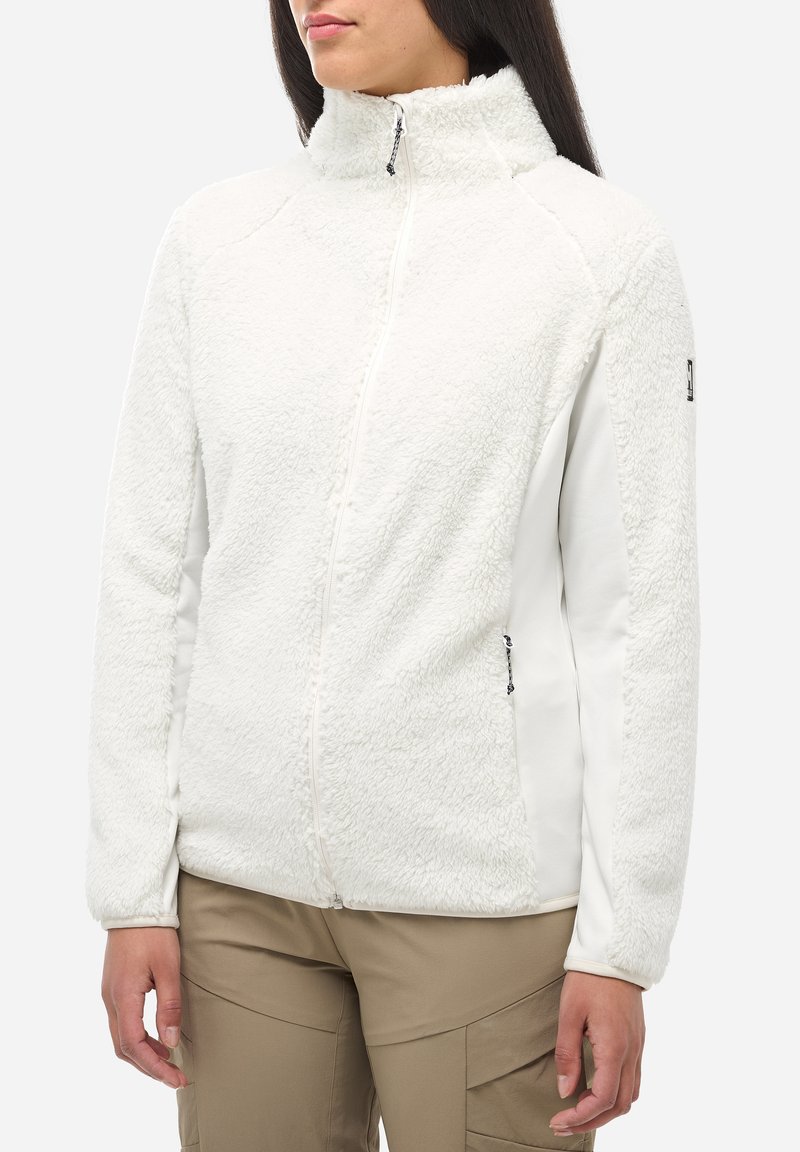 Femme portant une veste en polaire blanche zippée avec col haut et un pantalon cargo beige, se tenant devant un fond clair et uni.