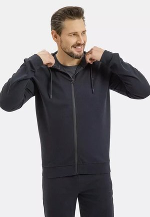 Mężczyzna w czarnej bluzie z kapturem na zamek i dopasowanych spodniach, regulujący sznurki od kaptura, uśmiechający się, na jasnoszarym tle.