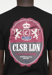 Fekete póló, hátán nagy, megkopott bordó és szürke grafikai dizájnnal, amely koronát, oroszlánokat és szöveges elemeket tartalmaz.