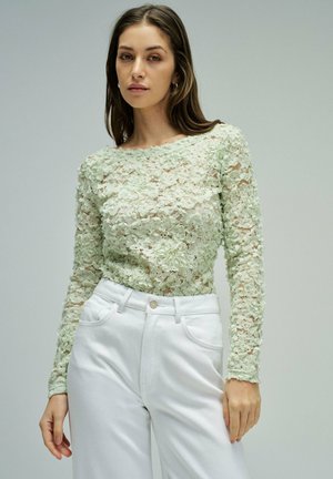 Femme portant un haut en dentelle florale vert menthe transparent à manches longues et un pantalon blanc taille haute, debout devant un fond uni.