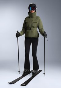 Zelena smučarska jakna z pasom, črne smučarske hlače, smučarske palice, goggles in čelada. Sleek dizajn z funkcionalnimi lastnostmi za smučanje.