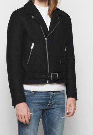 Personne portant une veste de motard noire avec des fermetures éclair argentées et une ceinture, un pull blanc et un jean bleu délavé, debout devant un fond uni.