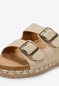 Sándalo de ante beige con dos hebillas ajustables, una plantilla acolchada y una suela texturizada envuelta en yute. Presenta un diseño plano y abierto.