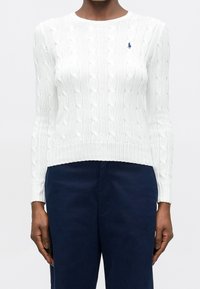 Personne portant un pull blanc cintré en maille torsadée avec un petit logo bleu marine sur la poitrine et un pantalon bleu foncé, debout devant un fond uni.