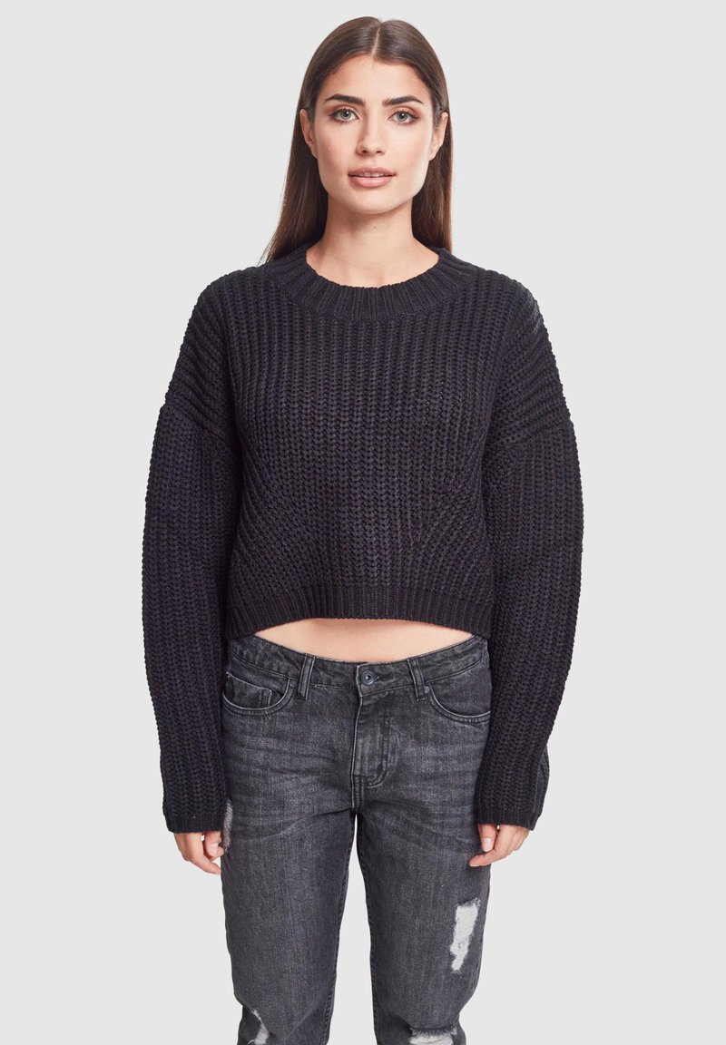Pull en tricot noir coupé avec un motif texturé et des manches larges, associé à un jean foncé détruit.