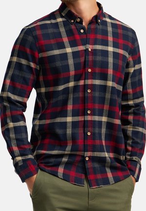 Hombre con camisa de cuadros en azul marino, rojo y beige, de manga larga y botones, y pantalones verde oliva, con las manos en los bolsillos.