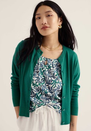 Jonge vrouw draagt een groene cardigan met knopen over een blauwgroene bloemen top en witte broek, met gouden creolen en gelaagde kettingen.